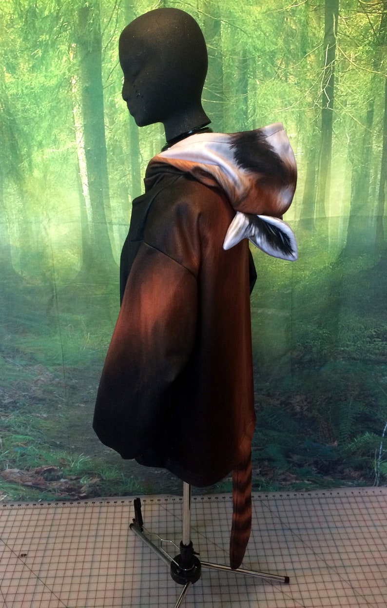 Puede incluir: Una sudadera marr&oacute;n y negra con un dise&ntilde;o de panda rojo. La sudadera tiene una capucha con una cabeza de panda rojo y una cola que cuelga de la espalda. La sudadera est&aacute; en un maniqu&iacute; contra un fondo de bosque verde.