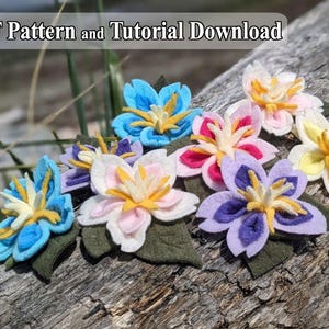May include: A collection of handcrafted felt flowers in various colors including blue, pink, white, yellow, and purple, each featuring layered petals and a central stamen, accompanied by dark green felt leaves and a PDF Pattern and Tutorial Download.