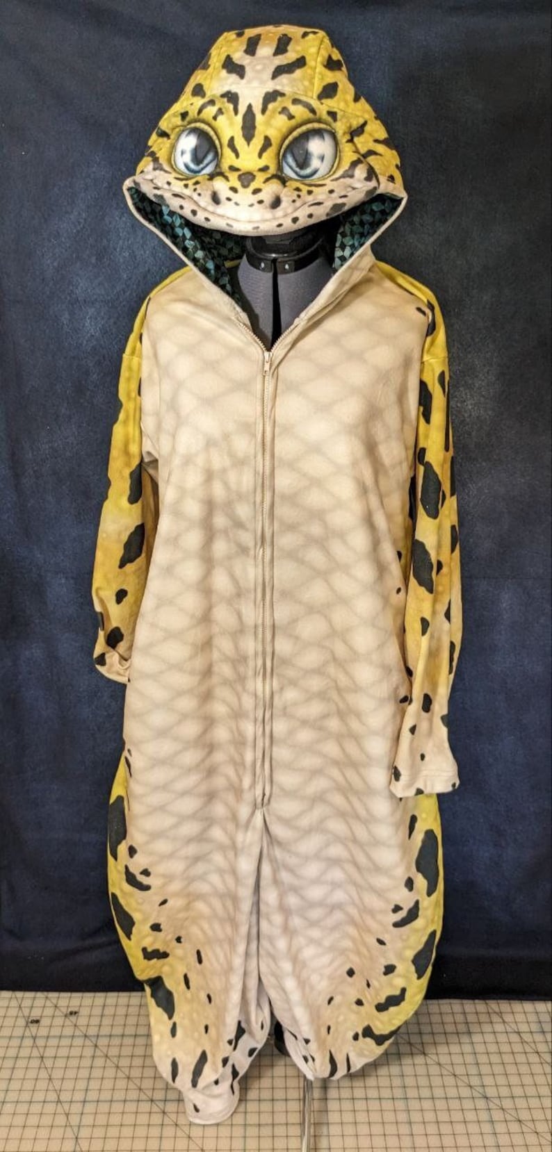 Puede incluir: Un mono beige de cuerpo entero con cremallera y estampado de leopardo amarillo y negro. El mono tiene una capucha con un dise&ntilde;o de cabeza de gecko en 3D.