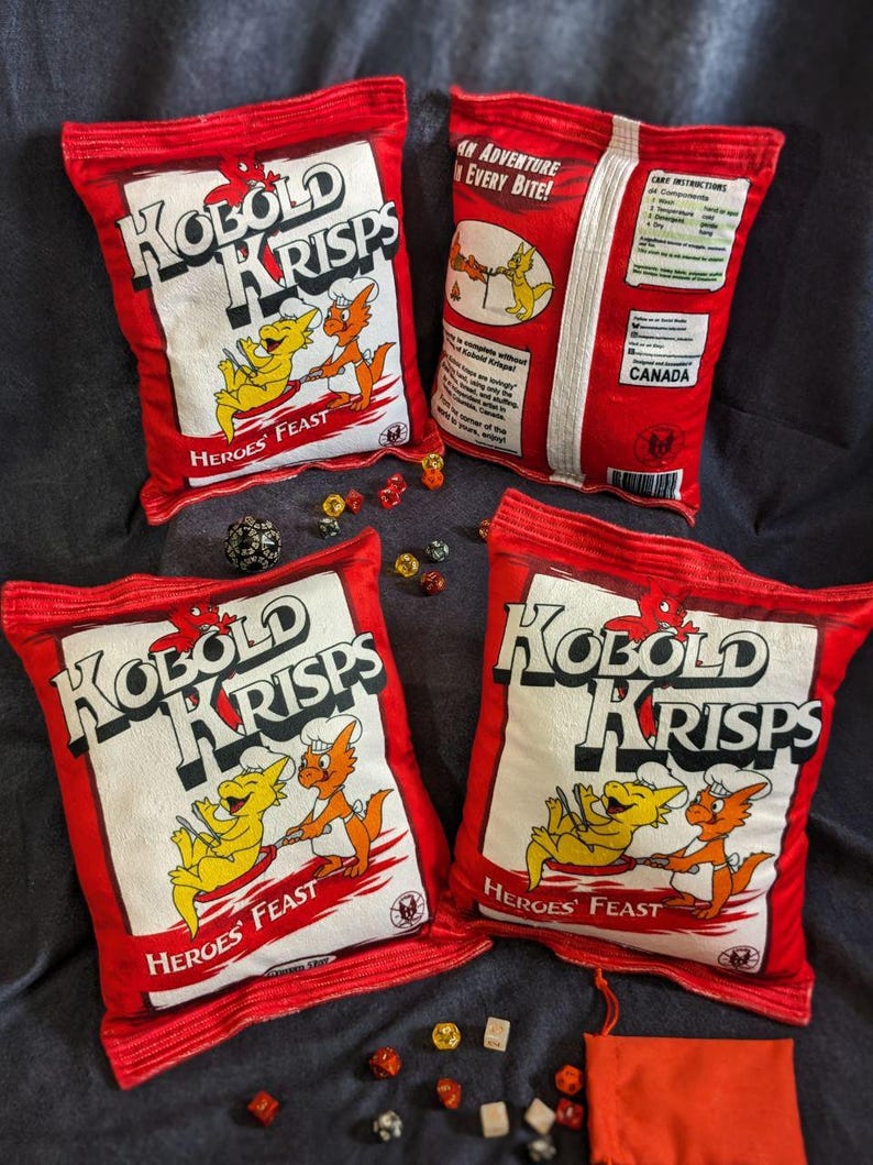 May include: Four red and white pillow-shaped chip bags with the words "Kobold Krisps" and "Heroes' Feast" printed on them. The bags are surrounded by dice and a red pouch.