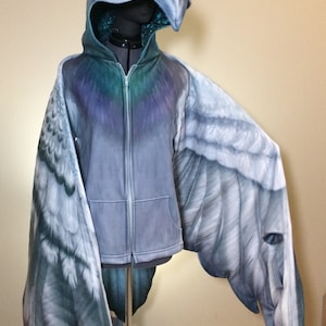 Puede incluir: Una sudadera con capucha gris con cierre de cremallera y alas adjuntas. Las alas est&aacute;n pintadas para parecer alas de p&aacute;jaro con un degradado de gris, blanco y azul.