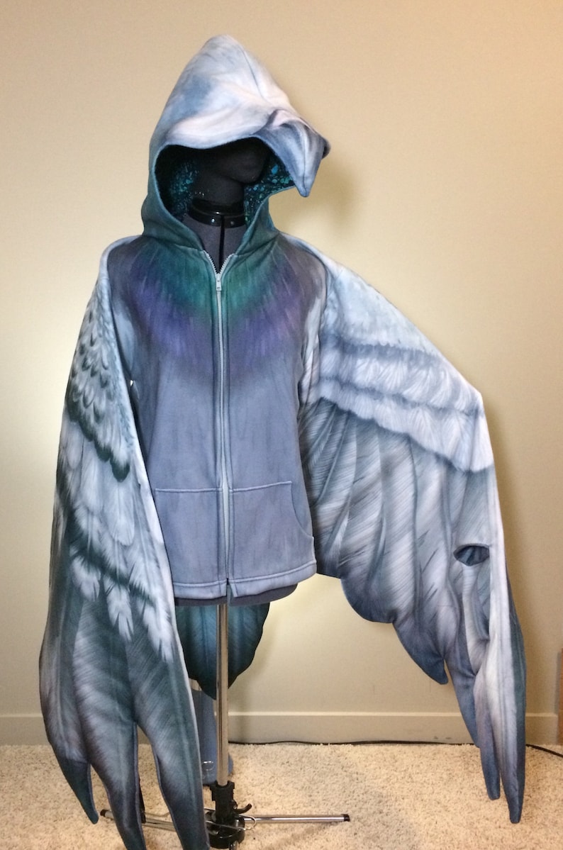 Puede incluir: Una sudadera con capucha gris con cierre de cremallera y alas adjuntas. Las alas est&aacute;n pintadas para parecer alas de p&aacute;jaro con un degradado de gris, blanco y azul.
