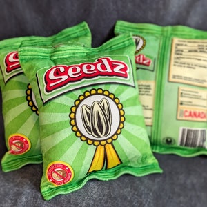 May include: Three green pillow-shaped bags with the word "Seedz" printed on them. The bags have a sunflower design and the text "Gluten Free" printed on them.