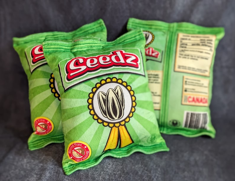 May include: Three green pillow-shaped bags with the word "Seedz" printed on them. The bags have a sunflower design and the text "Gluten Free" printed on them.
