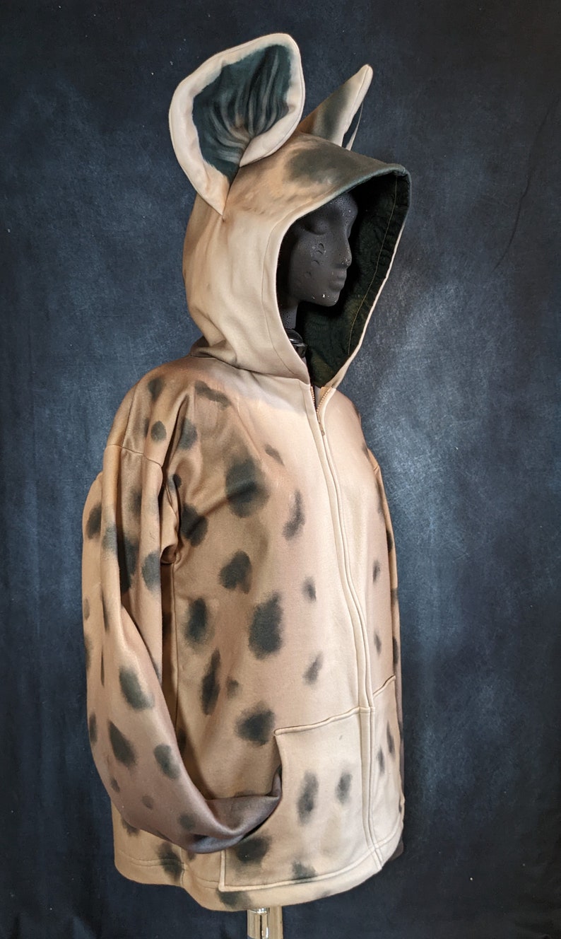 May include: A tan and black hooded sweatshirt with a spotted pattern and large ears. The sweatshirt has a zipper closure and a pocket on the front.