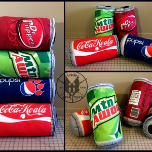 Puede incluir: Una pila de cuatro almohadas de peluche en forma de lata de refresco. Las latas son rojas, verdes, azules y rojas con las siguientes marcas: Dr Pepper, Mountain Dew, Pepsi y Coca-Cola.