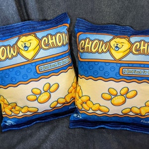 May include: Two blue and yellow plush pillows shaped like bags of chips. The pillows have the text "Chow Chow" and "A Good Snack For Good Dogs" printed on them.