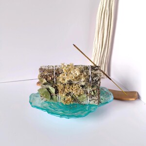 May include: A bundle of dried sage, tied with white string, adorned with dried flowers, and resting on a turquoise glass dish. An incense stick is lit, with a wooden holder. A macrame wall hanging is in the background.