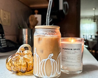 May include: A glass jar with a lid filled with iced coffee and a straw. The jar has a white pumpkin design. A glass candle jar with a white label that reads "Pumpkin Spice" is next to the coffee jar. A small glass pumpkin is in the foreground.