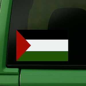 Può includere: Un adesivo rettangolare della bandiera palestinese, nei colori rosso, bianco, nero e verde, è attaccato al lunotto posteriore di un veicolo verde. L'adesivo è un rettangolo orizzontale con un triangolo rosso sul lato sinistro.