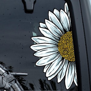 Può includere: Un adesivo a forma di girasole bianco e azzurro è applicato sul lunotto posteriore di un veicolo. L'adesivo è un design a mezzo girasole con un centro giallo ed è posizionato sul lato destro del finestrino. Il testo "Jen's Printing" è visibile sullo pneumatico.