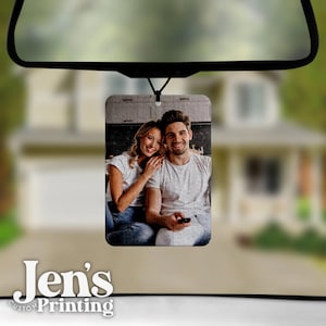 May include: A rectangular car air freshener hanging from a rearview mirror, featuring a black and white photograph of a smiling couple sitting together. The person on the left wears a white t-shirt and jeans, while the person on the right wears a white t-shirt.