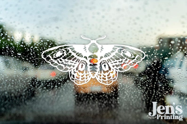 May include: A white moth-shaped decal is affixed to a window covered in raindrops. The moth has intricate wing patterns and a detailed body with orange, red, and black accents. The text "Jen's Custom Printing" is visible in the lower right corner.