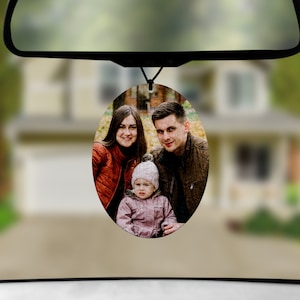 May include: A round car air freshener with a photo of a family of three. The photo shows a woman with long brown hair, a man with short brown hair, and a young child with blonde hair. The family is smiling and looking at the camera.