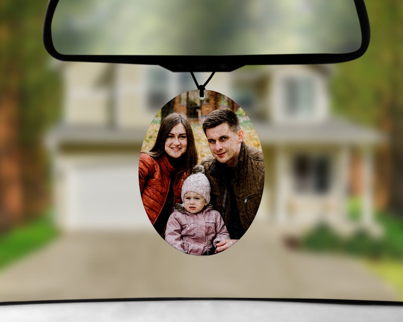 May include: A round car air freshener with a photo of a family of three. The photo shows a woman with long brown hair, a man with short brown hair, and a young child with blonde hair. The family is smiling and looking at the camera.