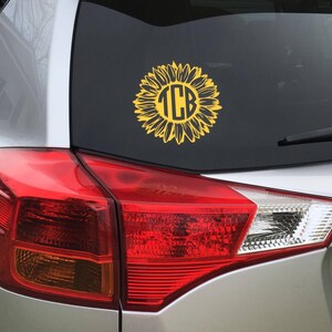 K&ouml;nnte beinhalten: Ein silbernes Auto mit einem gelben Sonnenblumen-Aufkleber auf der Heckscheibe. Die Sonnenblume hat einen schwarzen Kreis in der Mitte mit den Buchstaben "TCB" in Wei&szlig;.