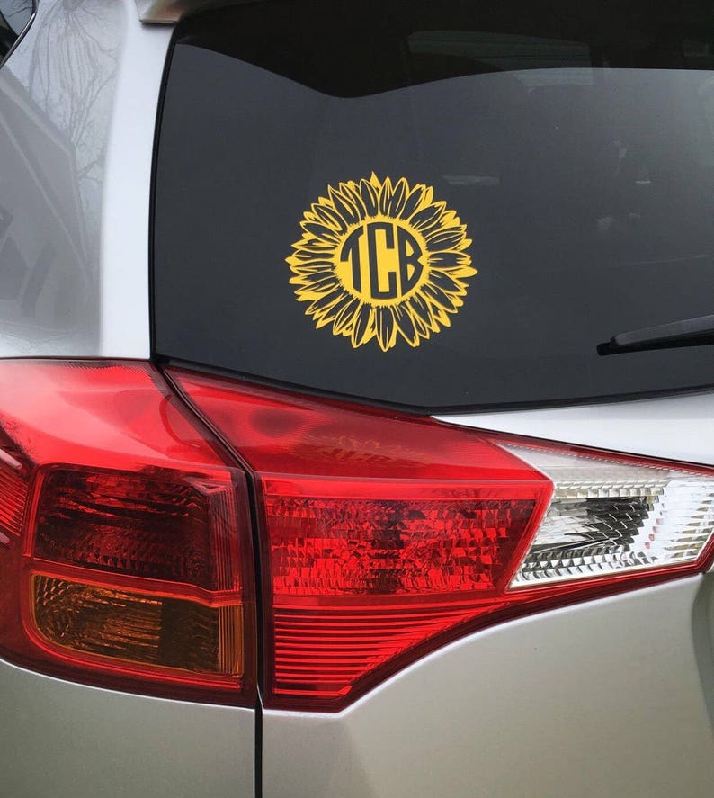 K&ouml;nnte beinhalten: Ein silbernes Auto mit einem gelben Sonnenblumen-Aufkleber auf der Heckscheibe. Die Sonnenblume hat einen schwarzen Kreis in der Mitte mit den Buchstaben "TCB" in Wei&szlig;.