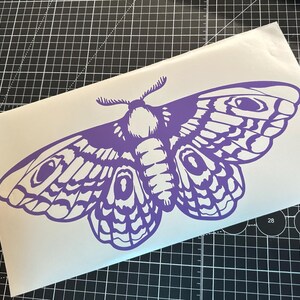 May include: A purple moth decal with intricate wing patterns and detailed body features. The decal is on a white background, with a black and gray grid in the background. The moth design is a striking, symmetrical illustration.