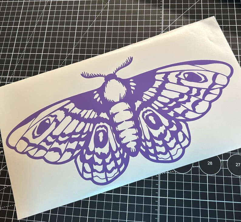 May include: A purple moth decal with intricate wing patterns and detailed body features. The decal is on a white background, with a black and gray grid in the background. The moth design is a striking, symmetrical illustration.