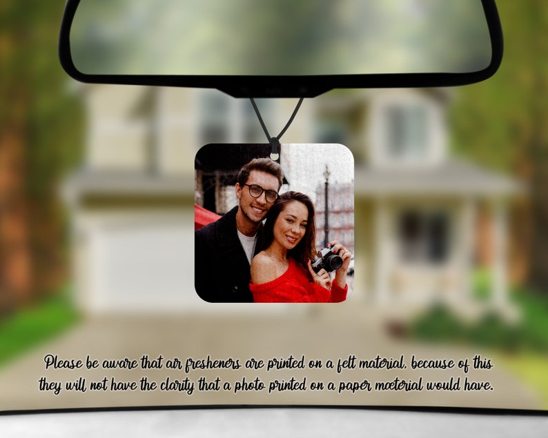 May include: A photo air freshener with a white background and a photo of a couple smiling. The man is wearing a black shirt and glasses. The woman is wearing a red shirt and holding a camera. The air freshener is hanging from a rearview mirror in a car.