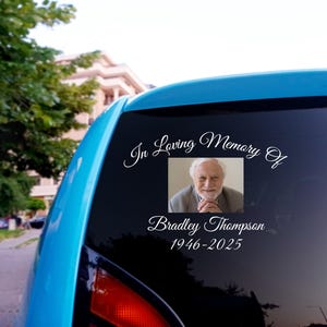 May include: A blue car with a white vinyl sticker on the back window. The sticker says "In Loving Memory Of" with a photo of a man and the text "Bradley Thompson 1946-2025".