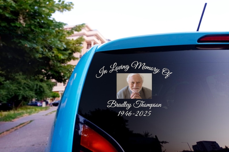 May include: A blue car with a white vinyl sticker on the back window. The sticker says "In Loving Memory Of" with a photo of a man and the text "Bradley Thompson 1946-2025".