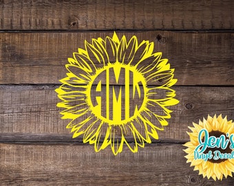 Puede incluir: Un girasol amarillo con un círculo en el centro que contiene las letras AMW. El girasol está sobre un fondo de madera marrón. Jen's Vinyl Decals está escrito en blanco sobre un girasol amarillo en la esquina inferior derecha de la imagen.