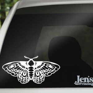 May include: White die-cut vinyl decal of a moth with detailed wing patterns, applied to a car window. The moth design is centered and prominent. The decal is a decorative element. The text "Jen's Custom Printing" is in the lower right corner.