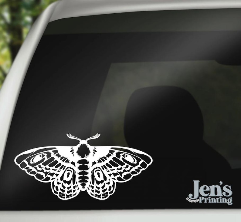 May include: White die-cut vinyl decal of a moth with detailed wing patterns, applied to a car window. The moth design is centered and prominent. The decal is a decorative element. The text "Jen's Custom Printing" is in the lower right corner.
