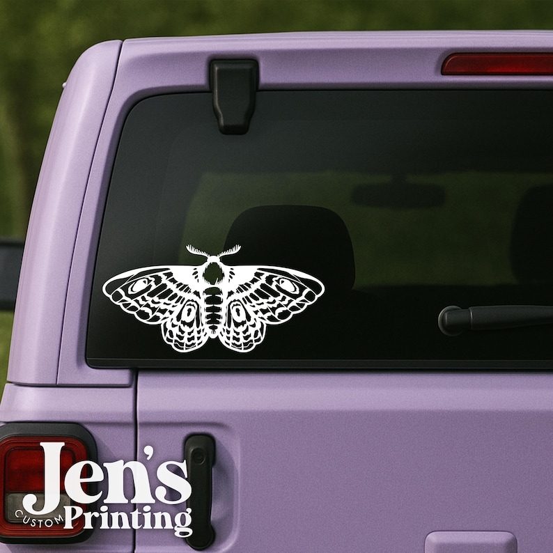 May include: White moth decal on a purple vehicle window. The decal features intricate wing details and antenna. The text "Jen's Custom Printing" is visible on the vehicle's rear. This decal is suitable for car enthusiasts and those who appreciate nature-inspired designs.