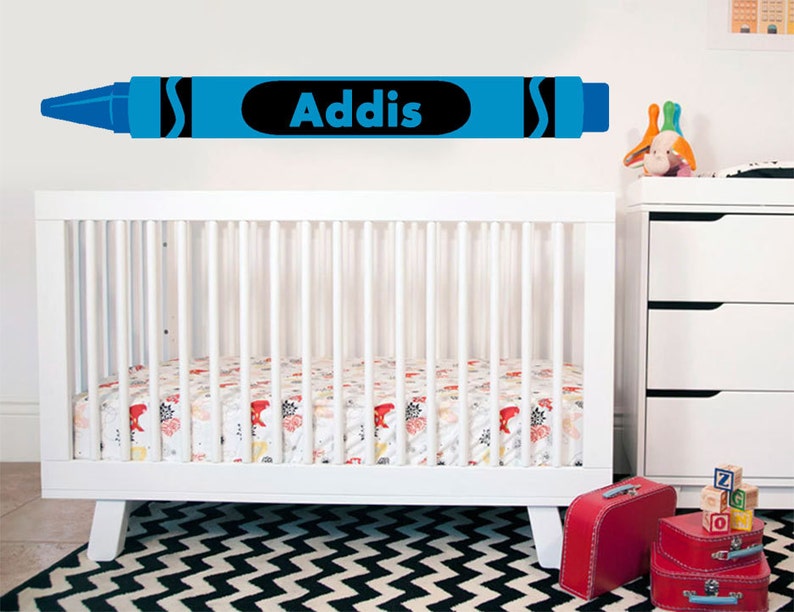 May include: A blue crayon-shaped wall decal with the name 'Addis' in black lettering is displayed above a white crib with a patterned mattress.  The crib is in a nursery setting with a white dresser and other baby items visible. The decal is a personalized nursery decoration.