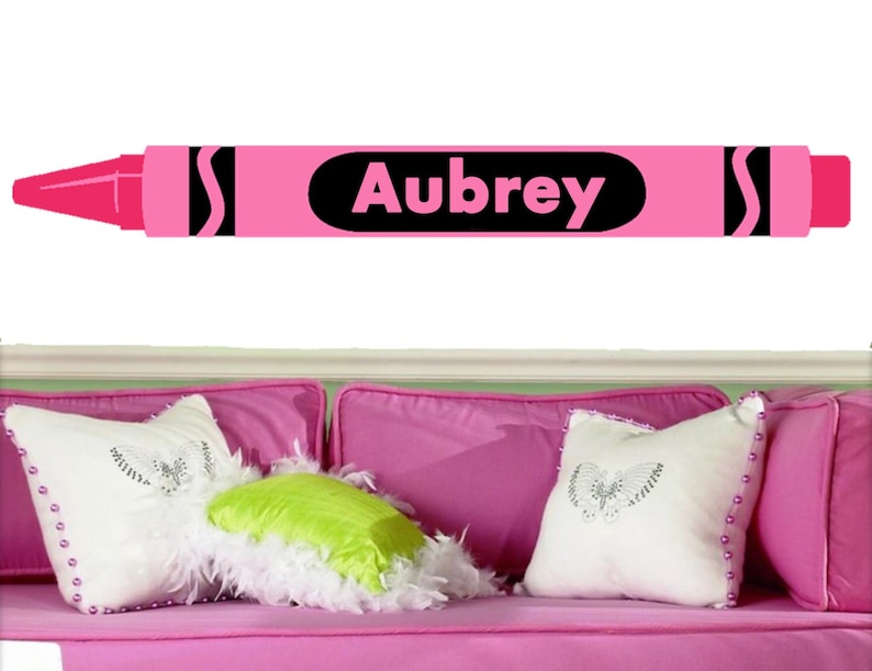 Puede incluir: Pegatina de pared de cray&oacute;n rosa con el nombre Aubrey escrito en negro. La pegatina tiene forma de cray&oacute;n con un cuerpo rosa y detalles negros.