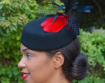 May include: A black felt hat with a red feather and black netting. The hat is worn on a person with dark hair.