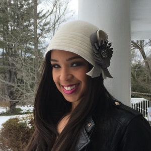 May include: A woman wearing a white felt cloche hat with a gray satin bow and a rhinestone embellishment. She is wearing a black leather jacket and has long dark hair.