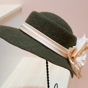 May include: A dark green felt hat with a wide brim and a white satin ribbon. The hat has a large, off-white fabric flower on the side.