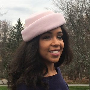 May include: A light pink cloche hat with a rounded crown and a slightly upturned brim. The hat is made of a soft, felt-like material. The hat is worn by a person with dark hair and a navy blue sweater.
