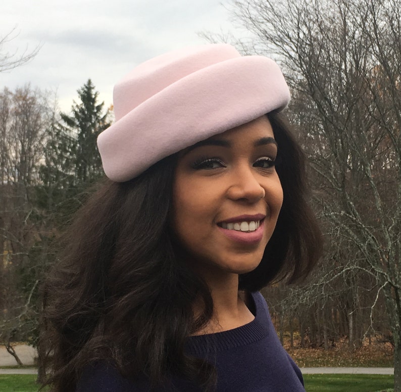 May include: A light pink cloche hat with a rounded crown and a slightly upturned brim. The hat is made of a soft, felt-like material. The hat is worn by a person with dark hair and a navy blue sweater.