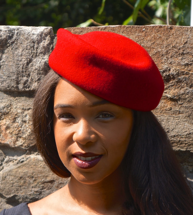 May include: A woman with long dark hair is wearing a red beret. The beret is tilted slightly to the side and covers part of her forehead. She is looking at the camera with a slight smile.