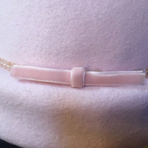 May include: A light pink hat with a soft, felt-like texture. The hat is adorned with a pale pink velvet ribbon and a string of small, clear beads. The hat's brim is slightly curved, and the overall design is simple and elegant.