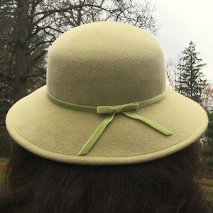 May include: A light green cloche hat with a rounded crown and a wide brim. The hat features a light green ribbon around the crown and a bow on the front. The hat is made of a soft, felt-like material.