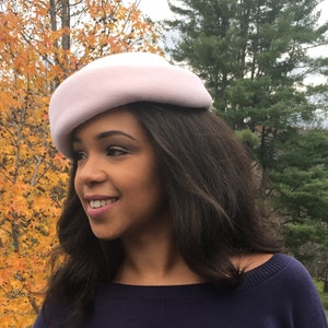 May include: A light pink beret-style hat. The hat is made of a soft material and has a rounded crown. The hat is worn by a person with dark hair and a navy blue top. The background features fall foliage.