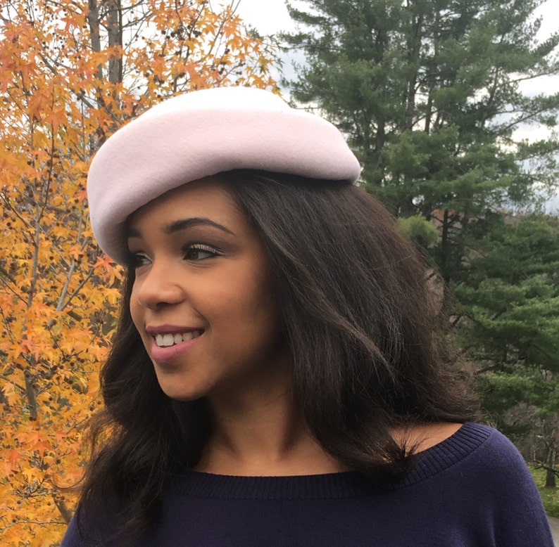 May include: A light pink beret-style hat. The hat is made of a soft material and has a rounded crown. The hat is worn by a person with dark hair and a navy blue top. The background features fall foliage.