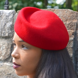 May include: A red felt beret hat. The hat is round and has a slight curve to it. The hat is worn on a person's head.