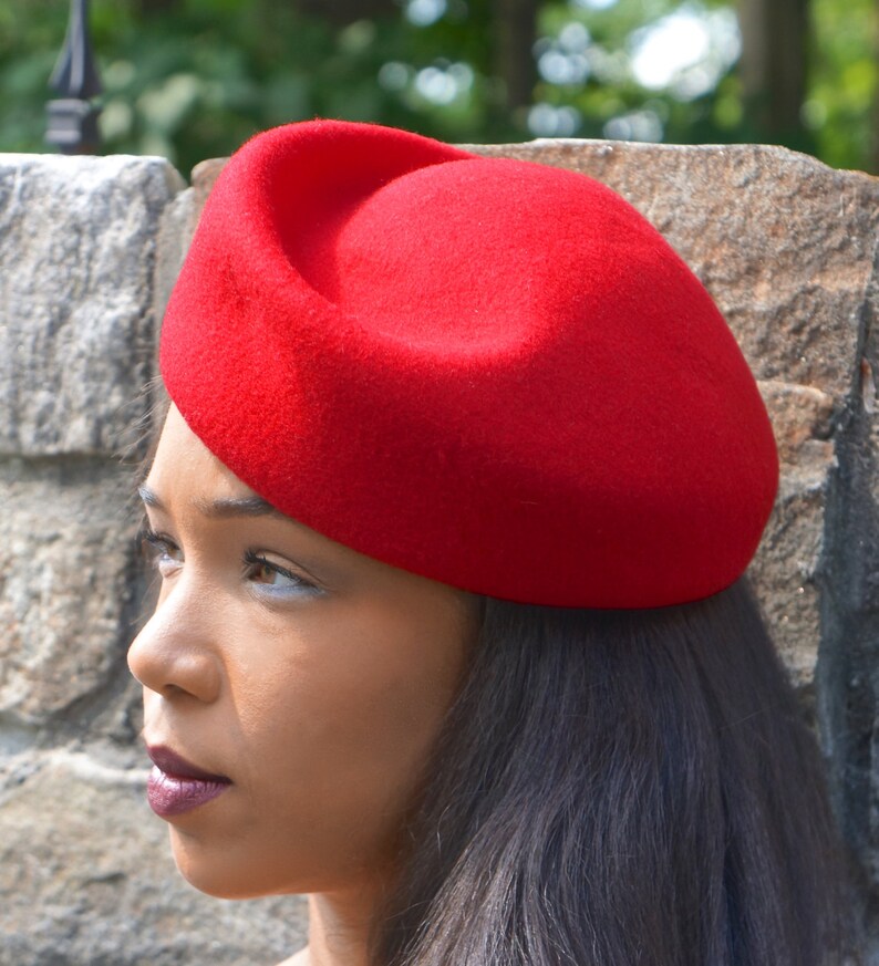 May include: A red felt beret hat. The hat is round and has a slight curve to it. The hat is worn on a person's head.