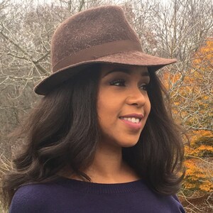May include: A woman with long brown hair is wearing a brown felt fedora hat with a brown ribbon. She is wearing a dark blue sweater and is standing in front of a background of trees with fall foliage.