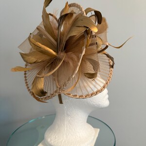 May include: A gold-toned fascinator with a circular base and a large, feathery floral design. The headpiece features layers of mesh, feathers, and decorative elements, creating a sophisticated and elegant look. The headpiece is displayed on a white mannequin head.