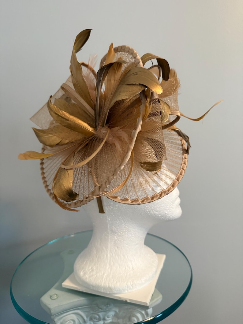 May include: A gold-toned fascinator with a circular base and a large, feathery floral design. The headpiece features layers of mesh, feathers, and decorative elements, creating a sophisticated and elegant look. The headpiece is displayed on a white mannequin head.