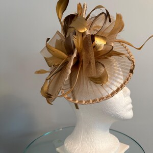 May include: A gold-toned fascinator with a circular base and a decorative arrangement of feathers and netting. The headpiece is displayed on a white mannequin head, showcasing its design and structure. The feathers have a metallic sheen.