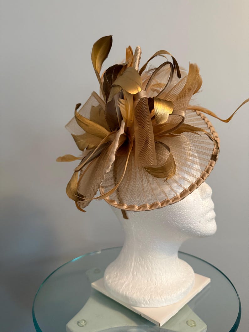 May include: A gold-toned fascinator with a circular base and a decorative arrangement of feathers and netting. The headpiece is displayed on a white mannequin head, showcasing its design and structure. The feathers have a metallic sheen.