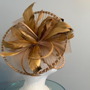 May include: A gold-colored fascinator with a circular base and layered design. The headpiece features large, stylized feathers and a sheer, net-like material. The hat is displayed on a white mannequin head.